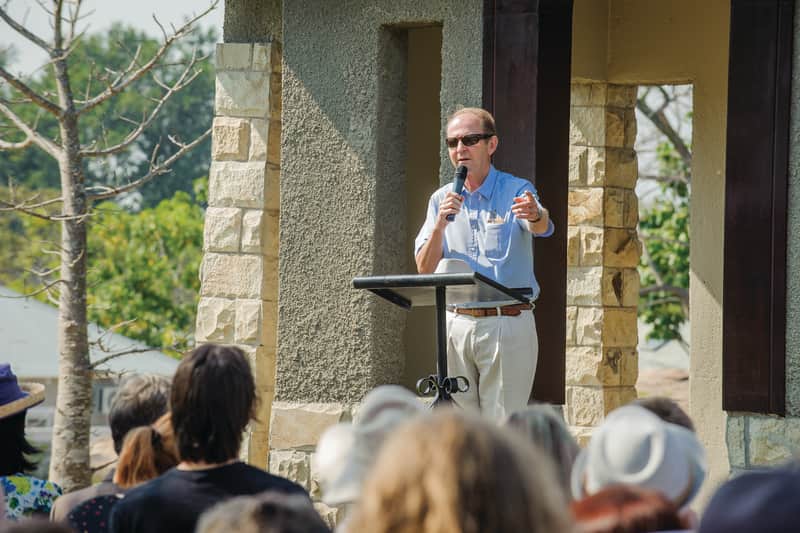 A speaker with a microphone addresses the gathered family from a stone pavilion at the dedication service
