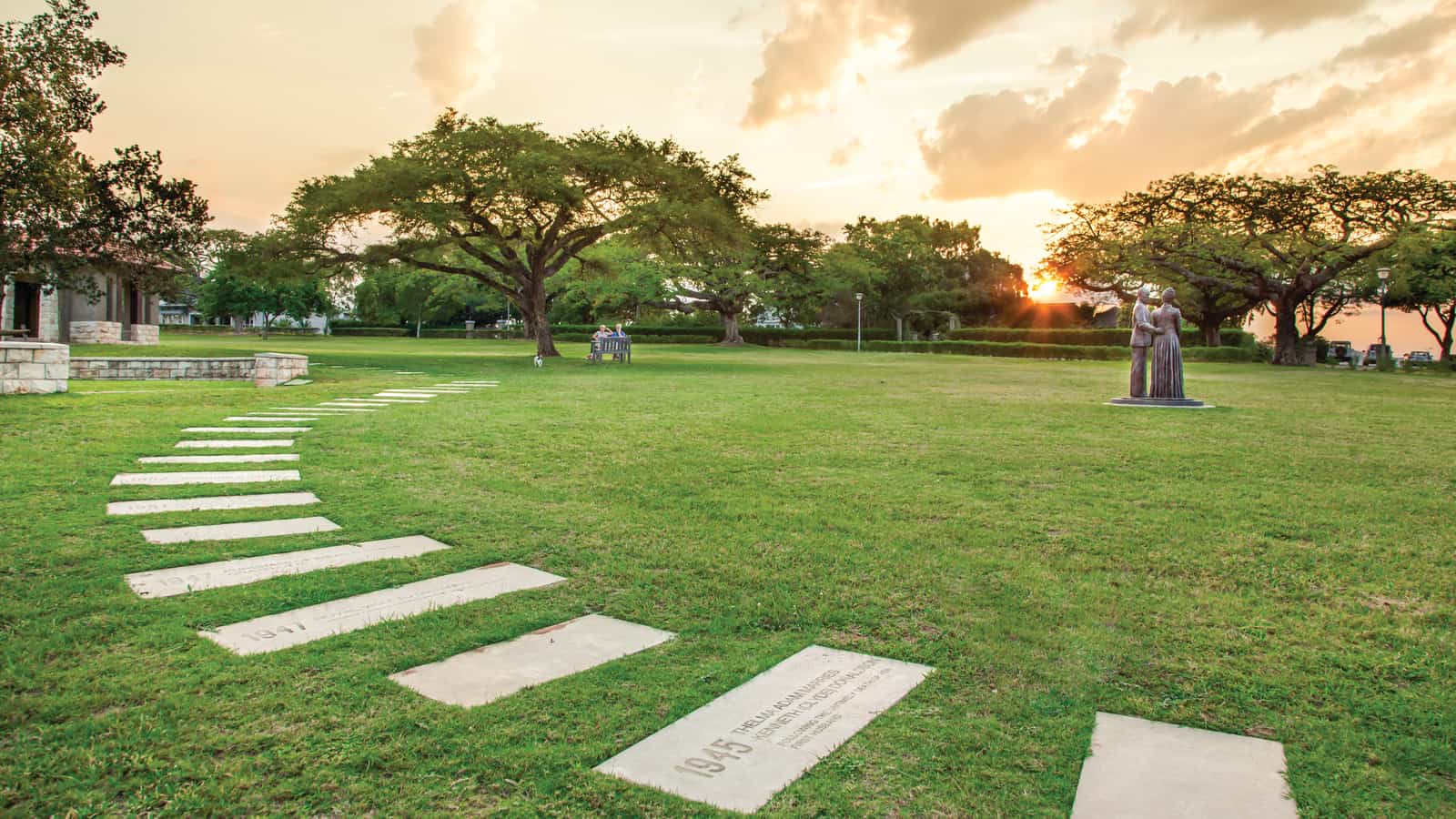 The Halls Heritage Park at Mataffin at sunset: a bronze statue of the founding couple on a broad lawn, a pathway of dated milestone stones curving across the grass beneath wide trees