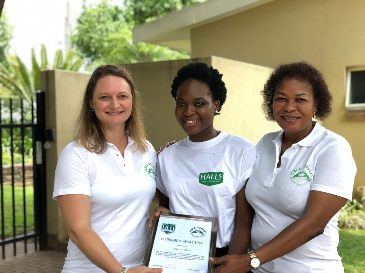 Three women in Halls and Lula Sandla shirts presenting a framed certificate of appreciation