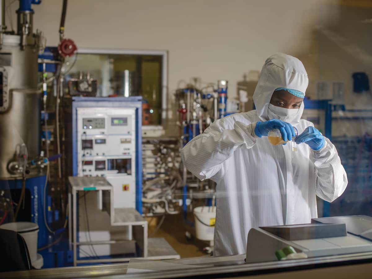 A scientist in a cleanroom suit and gloves decants a sample from a flask at a laboratory bench, bioreactors behind the glass