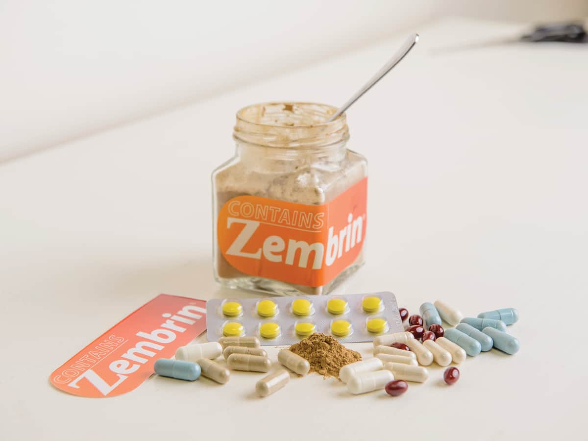 A jar of Zembrin powder with a spoon, surrounded by capsules, tablets and loose extract on a white table